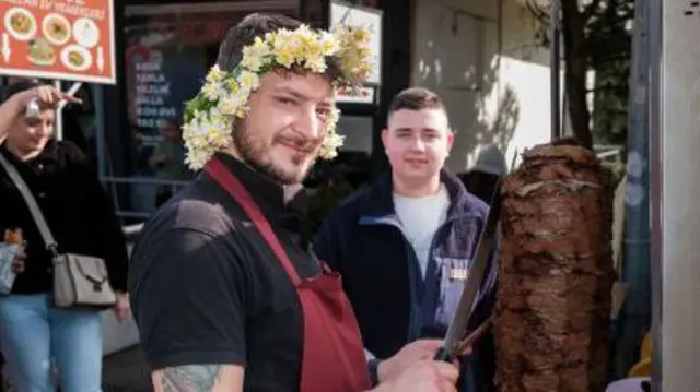 Мастер донера в короне из нарциссов стоит рядом с куском мяса на вертеле и ножами в руках.