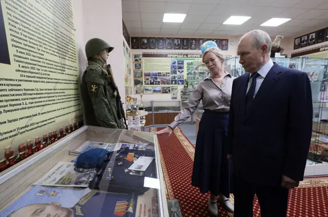 Vladimir Putin mira una vitrina donde están presentados artículos de soldados en una especie de museo militar, mientras una mujer con una falda larga, le explica al líder lo que ve.