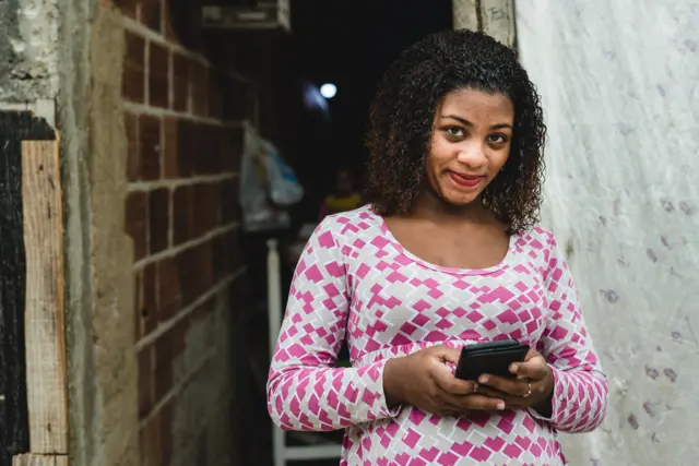 Mulher jovembrazino 7baixa renda segurando um celular e sorrindo timidamente para a câmera