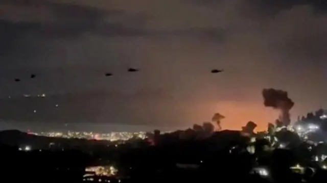 Imagem estática ligeiramente desfocada de um vídeo mostrando pelo menos 5 helicópteros sobrevoando Caracas, e colunas de fumaça podem ser vistas subindo do solo.