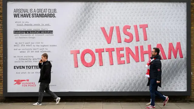 Le panneau Visit Tottenham se trouve à l'ombre de l'Emirates Stadium d'Arsenal. Deux supporters d'Arsenal passent devant un panneau sur lequel on peut lire « Visit Tottenham » en lettres rouges sur fond blanc. En lettres plus petites, on peut lire : "Arsenal est un grand club. Nous avons des normes"