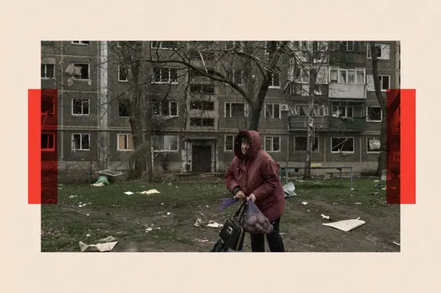 Жінка з сіткою картоплі та сумочкою на фоні будинку зі слідами обстрілів