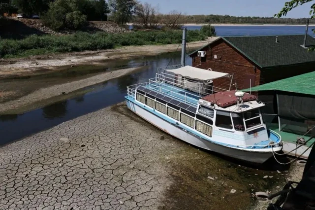 Обмелевшая река, ушедшая вода открыла растрескавшееся дно. На берегу стоит кораблик: судя по всему, раньше он был в воде, теперь стоит на грунте.