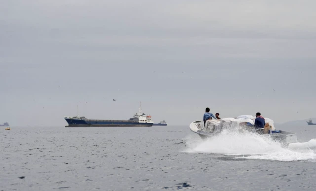 Üç kişinin içinde olduğu bir deniz motoru hızla ilerliyor. Arkada büyük bir tanker var.