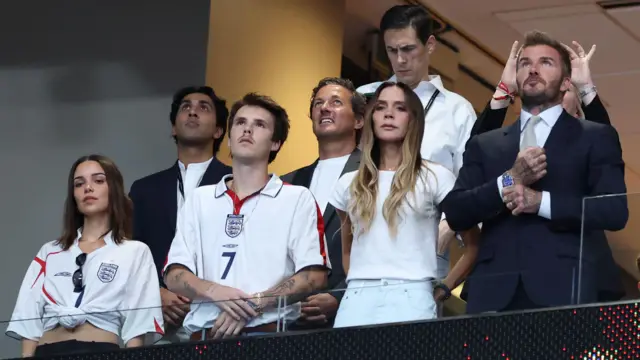 A picture of Jackie Apostel, Cruz Beckham, Victoria Beckham, David Beckham and Ken Paves in a box at a football match during the World Cup between England and Argentina. The group, dressed in a mixture of England shits and formal clothing, appear to be looking up at a screen, which is out of shot.