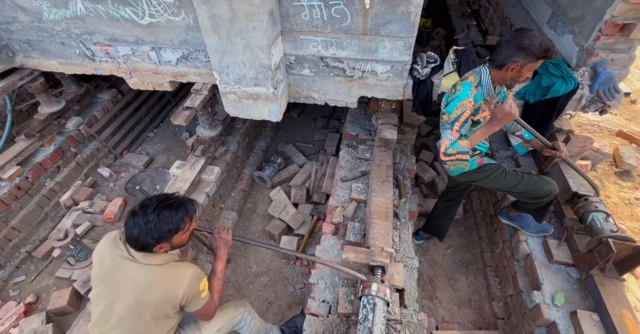 ਨੀਹਾਂ 'ਤੇ ਜੈੱਕ ਲਗਾਉਂਦਾ ਅਤੇ ਮਕਾਨ ਨੂੰ ਅੱਗੇ ਖਿਸਕਾਉਂਦੇ ਹੋਏ 