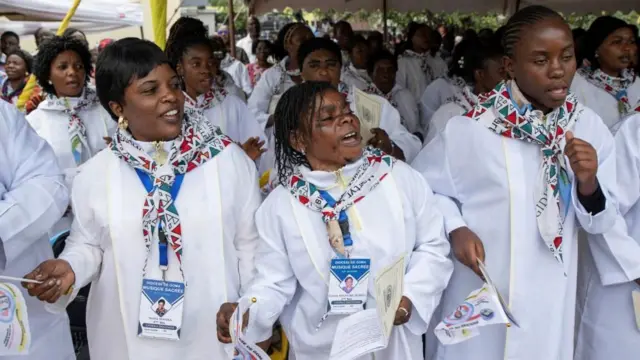 Abagize korali gatolika barimo kuririmba mu misa yo kwizihiza kugirwa Umuhire kwa Floribert. Bambaye amakanzu y'umweru, barimo kuririmba ndetse barimo kureba iburyo bw'iyi foto.