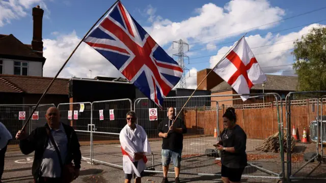 Manifestantes sostienen banderas con la Union Jack y la Cruz de San Jorge afuera del edificio de un hotel rodeado por una valla de metal.
