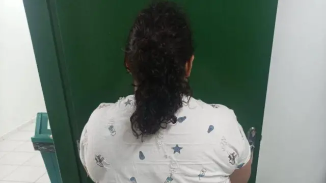 Koyu yeşil bir kapının önünde duran, beyaz bluz giymiş ve koyu renk saçlarını at kuyruğu yapmış bir kadının arkadan görünümü.