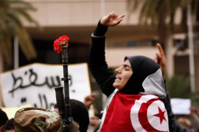 Gabar Tuniisiyaan ah oo garabkeeda ka muuqdo qori afka kaga gufaysan yahay ubax, xili lagu jiro mudaharaadyo looga soo horjeedo dowlada Tunisia, Janaayo 2011.