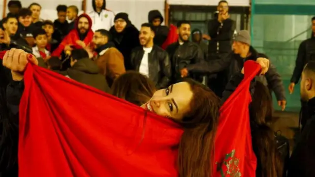 A woman with long hair posing with a Moroccan flag. She is smiling and there is a jubilant crowd of men in the background.