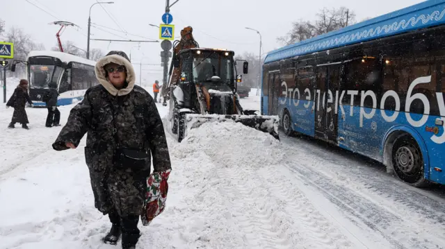 Женщина идет по снегу, за ее спиной снегоуборочная машина, справа актобус с надписью «Это электробус», позади слева трамвай, перед которым перебегают дорогу два человека