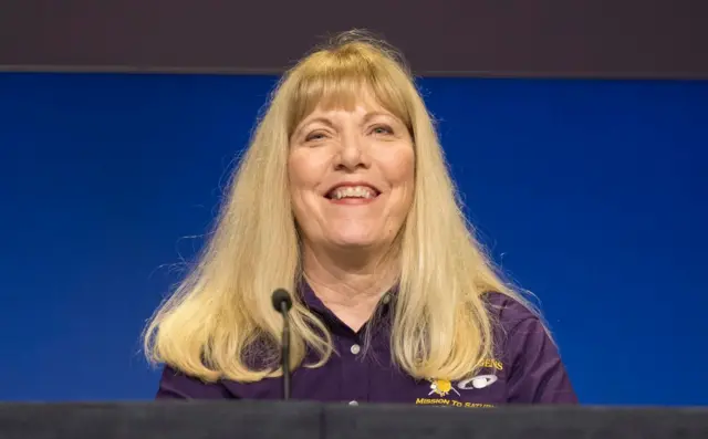 A photo of a woman with long blonde hair smiling as she sits behind a microphone at a table, wearing a purple shirt with a space logo