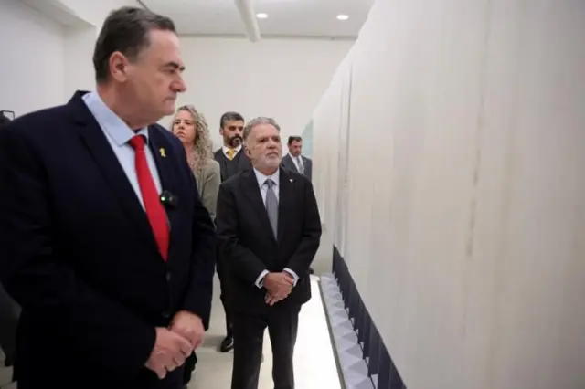 Em destaque, dois homens engravatados e braços contidos observam parede de museu