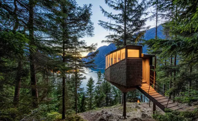 Una cabaña rectangular se erige sobre una loma que da a un lago y dos montañas que surgen a lado y lado.