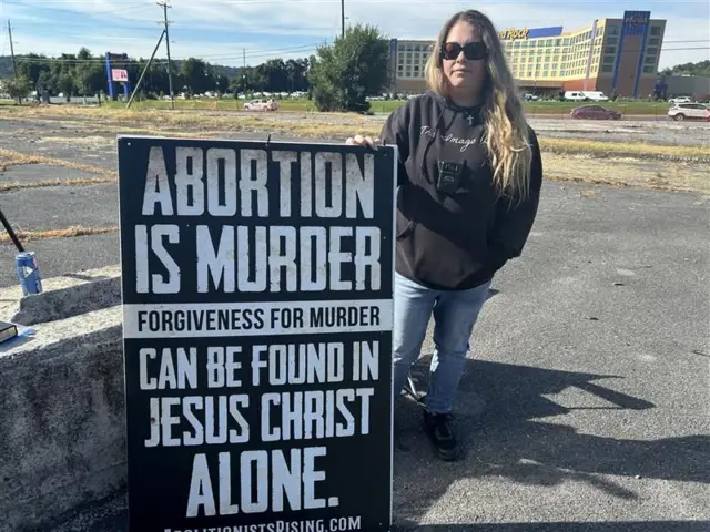 Uma mulher está em pé em um estacionamento segurando um cartaz que diz: “Aborto é assassinato. O perdão pelo assassinato pode ser encontrado somente em Jesus Cristo”.
