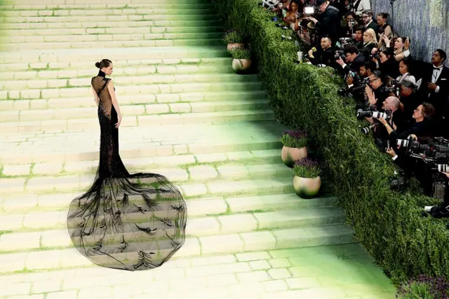Una persona posa sola en una amplia escalinata verde durante un evento de gala, con un vestido negro con larga cola transparente, mientras fotógrafos capturan imágenes desde un lateral.