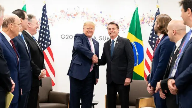 Donald Trump e Jair Bolsonaro apertando as mãos com bandeiras dos Estados Unidos e Brasil ao fundo e cercadosmines onabetassessores