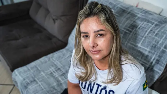 کلودیا با تی‌شرتی خاکستری روی مبل نشسته و به دوربین نگاه می‌کند. موهای بلوندش روی شانه‌هایش افتاده است.