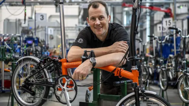 Will Butler-Adams, presidente executivo da Brompton Bicycle, com uma camisa polo preta apoiado em uma bicicleta laranja