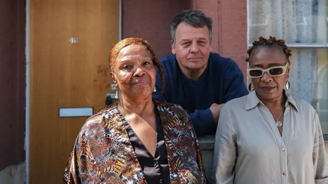 Três indivíduos estão em frente a um edifício de textura marrom-avermelhada. A pessoa à esquerda usa uma jaqueta estampada com várias cores e desenhos, incluindo tons de marrom, preto e laranja. A pessoa no meio usa uma camisa de manga comprida azul-escura e se inclina ligeiramente contra a parede. A pessoa à direita usa uma camisa de botão de cor clara e um colar com um pingente circular. Atrás deles, há uma porta de madeira marrom-alaranjada marcada com o número "46" e um escaninho de correspondência abaixo dela. À direita da porta há uma janela com cortinas de renda.