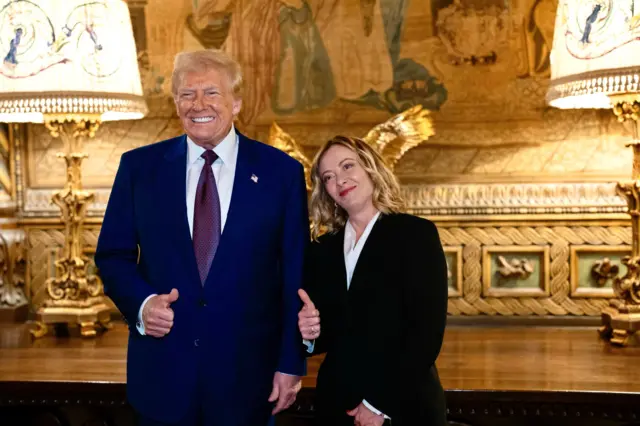 Trump e Meloni em salão posando para foto; ambos sorriem e fazem sinal com polegar