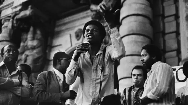 Homem negro ao centro falando em um microfone