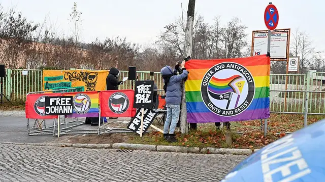 Демонстранты протестуют перед началом суда 25 ноября 2025 года. На плакатах белым по черному написано «Свободу Йохану». Человек, стоящий спиной к камере в голубой куртке и капюшоне, вешает радужный флаг, на котором написано «Квир и антифашист», «Объединяться и противостоять»