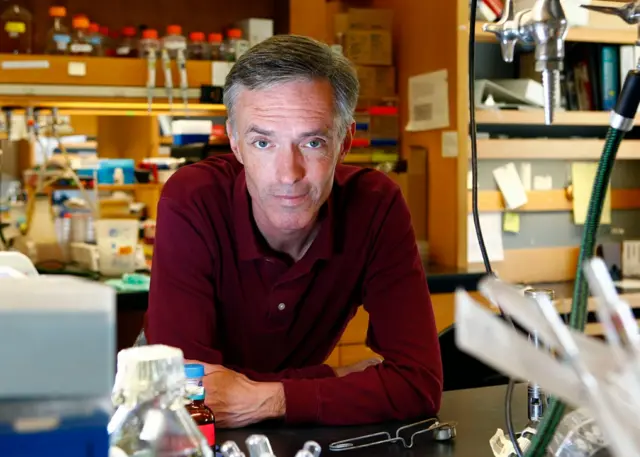 پروفسور کالینز روی میز آزمایشگاهش خم شده است و پیراهنی زرشکی به تن دارد. در پس‌زمینه، مجموعه‌ای از تجهیزات علمی دیده می‌شود که در حالت تار قرار دارند.