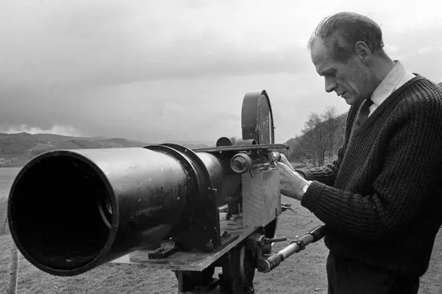 在一張黑白照片中，一名男子正在調整長鏡頭相機的設定。遠處尼斯湖的湖水清晰可見。