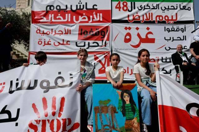 أطفال يجلسون حول لافتات، يعرض بعضها رسائل تدعو إلى إنهاء حرب غزة خلال مسيرة يوم الأرض