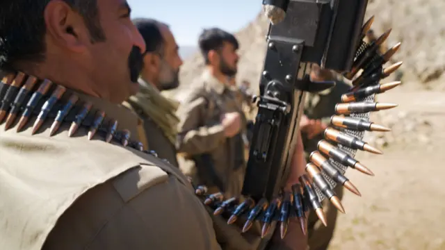 سه جنگجوی کرد ایرانی ایستاده‌اند و به سمت راست نگاه می‌کنند. نزدیک‌ترین آنها یک سلاح در دست دارد و یک کمربند مردمی به گردنش بسته است.