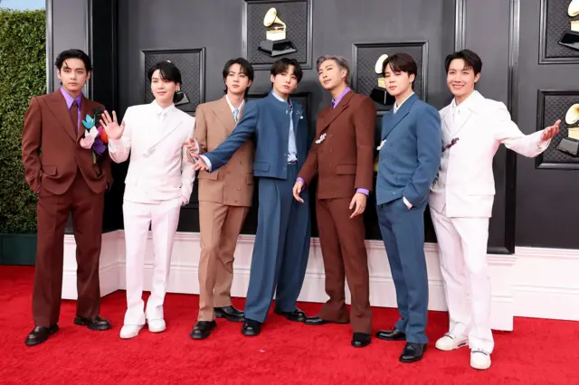 Los miembros de BTS posan en fila, vestidos con trajes, frente a un fondo con el logotipo de los premios Grammy durante la alfombra roja de los Grammy de 2022.