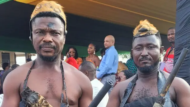 Deux porte-flingues royaux, vêtus de chapeaux et de vêtements traditionnels, se tiennent debout et portent des armes de cérémonie.