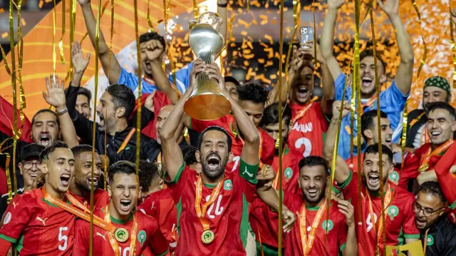 Le capitaine marocain Mohamed Rabie Hrimat brandit le trophée incurvé du Championnat d'Afrique des Nations, en forme de vase, entouré de ses coéquipiers en maillots rouges, médailles autour du cou et confettis orange flottant au vent.