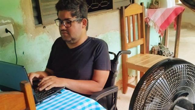 Guilherme sentado na cadeira de rodas diante de laptop e perto de ventilador