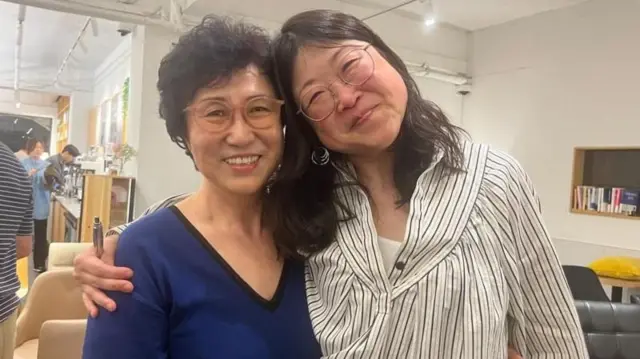 Mãe e filha abraçadas no interior de uma sala, olhando para a câmera