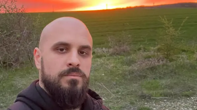 Muškarac bez kose sa bradom gleda u kameru tokom zalaska sunca