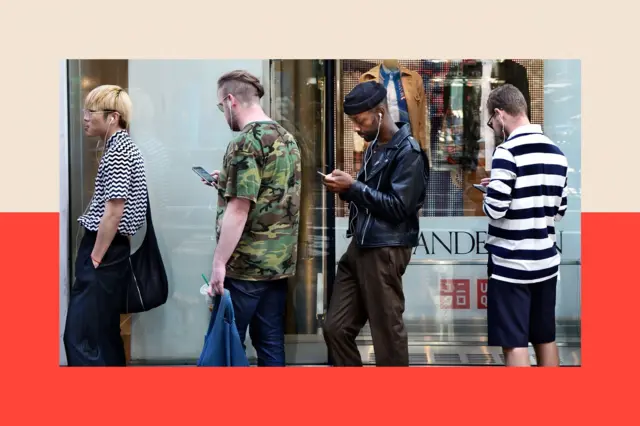 Four men stand in a queue, looking at their smartphones.