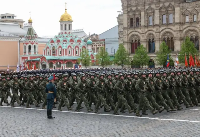 Россия ҳарбий хизматчилари тўқ яшил форма кийиб, дубулғаларига кўзойнак тақилган ҳолда ва қўлларида қурол ушлаб саф тортиб юриш қилмоқда. Уларнинг ортида кўк-яшил, қизил ва оқ рангларга бўялган, олтин гумбазли Қозон бош черкови кўринади.