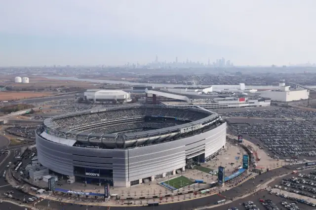 MetLife Stadyumu'nun kuşbakışı görüntüsü, devasa oval yapısını gösteriyor. Cephede "METLIFE STADIUM" yazısı görülüyor. Mekan, yoğun bir şekilde dolu otoparklarla çevrili. Uzakta, New York şehrinin ikonik silüeti ufukta yükseliyor.
