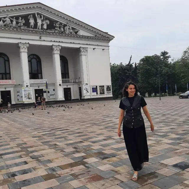 Дівчина у чорній сукні і шкіряному жакеті стоїть на площі перед тетаром