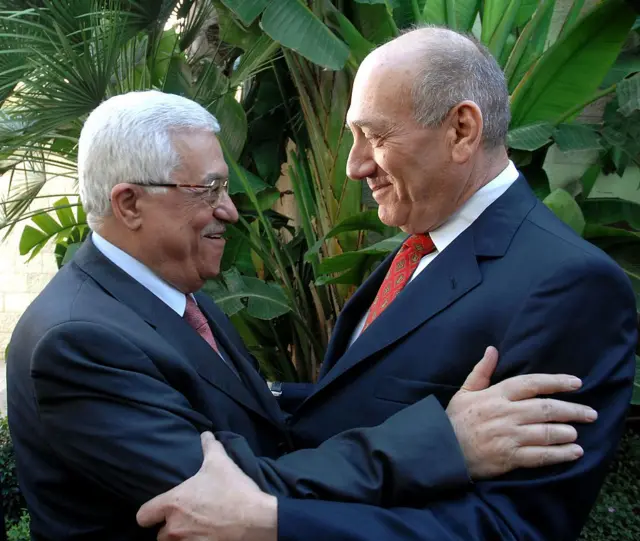 Mahmud Abás y Ehud Olmert sonríen y se abrazan. Ambos van vestidos con trajes de chaqueta azul marino, corbata roja y camisa blanca. Abás lleva gafas y tiene el pelo blanco y Olmert es calvo.