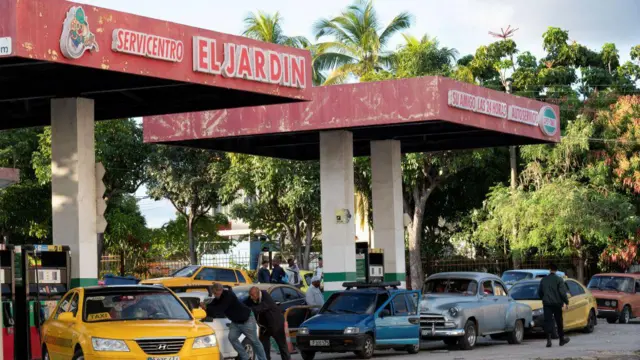 Una fila en una gaslinera en cuba. Hay varios autos, el establecimiento se ve viejo y en mal estado. 