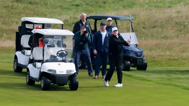 Дональд Трамп, одягнений у синій светр, штани та білу кепку збірної США, робить удар по м'ячу для гольфу на фервеї, поки інші члени групи спостерігають поруч із гольф-баггі.