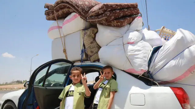 Dva dečaka gestikuliraju dok sirijska izbeglička porodica ulazi u automobil natovaren stvarima
