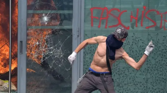 Manifestante sem camisa,nordeste futebol bet apostarosto coberto e boné, atira objetonordeste futebol bet apostafrente a uma fachada com vidros quebrados, refletindo um incêndio