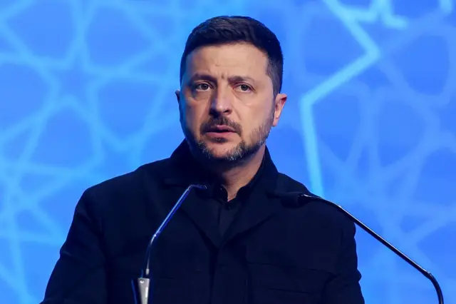 Зеленський у темному, напівофіційному костюмі виступає за трибуною з двома мікрофонами на тлі синього екрана з геометричним візерунком.