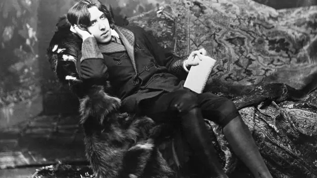 Черно-белая фотография Оскара Уайльда, он вальяжно и задумчиво сидит на диване и держит в руке блокнот.