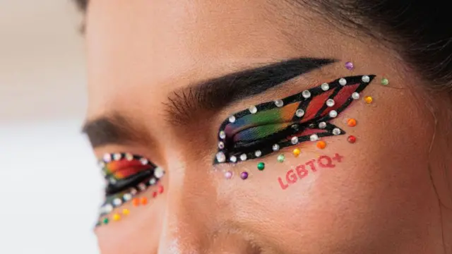 Mulher com sombra e maquiagem da cor do arco-íris, ostenta as letras LGBTQ +aposta agoravermelho abaixo dos olhos fechados